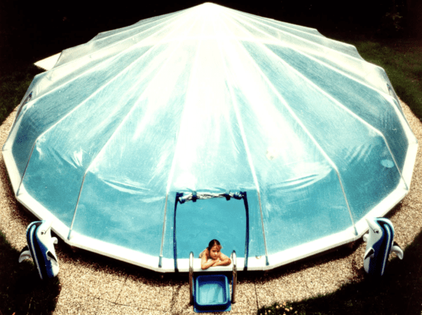 A child sits on a blue pool deck beside an oval swimming pool covered with a translucent Round Above Ground Sun Dome For Steel Wall Pools, viewed from above.